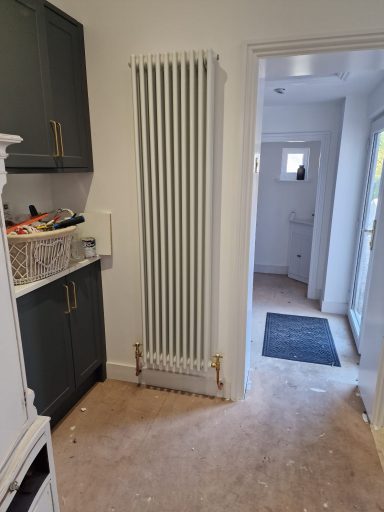 Narrow white vertical radiator mounted on a wall in a light hallway with a doorway.