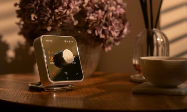 A modern device with a large dial, placed on a wooden table near flowers and a bowl.