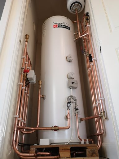 A vertical white hot water cylinder with copper pipes and fittings.